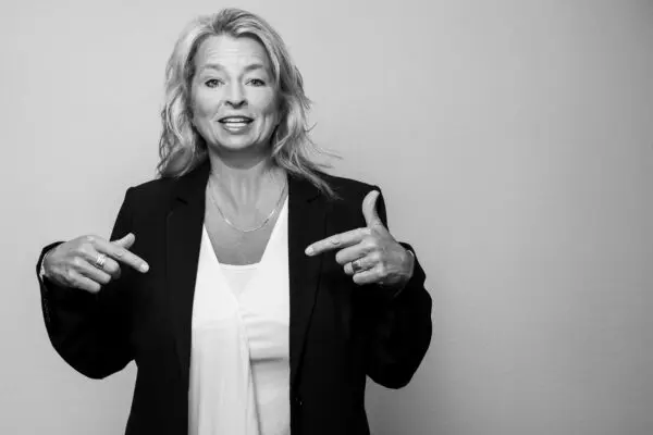 Woman in blazer pointing at herself, smiling.